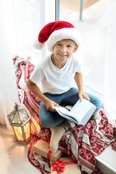 Cute Little Child In Santa Hat Reading Christmas Story While Sitting On Windowsill At Home