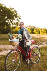 Young smiling girl on red bicycle with wildflowers and fruits in bicycle basket happily looking in camera in city park