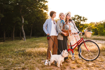 Beautiful girls with bicycle and wood basket in hands dreamily looking aside while happily spending time with little dog in park