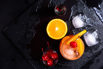 Tequila Sunrise cocktail, orange, ice cubes, maraschino cherries and jigger on a wet black slate...