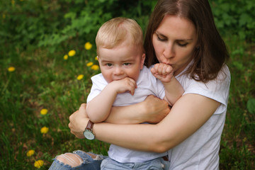 Young pregnant girl sits on lawn in city park. Mother hugs and kisses her son's hand. Little boy bites his hand. Happy motherhood