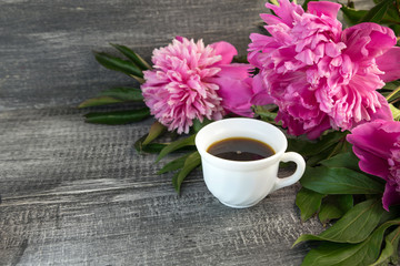 Obraz na płótnie Canvas White cup of coffee with bouquet of large bright pink peonies on a grey wood table with copy space.