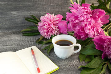 Obraz na płótnie Canvas White cup of coffee with bouquet of large bright pink peonies with notebook and red pen on a grey wood table.