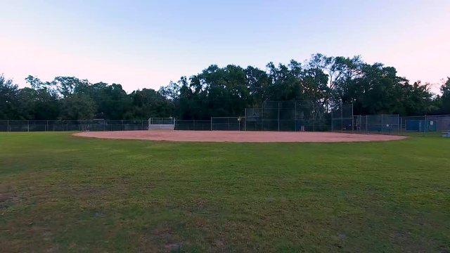 This clip is an aerial, drone fly over of a baseball field in a park. The drone flies over the field, then ascends over the fence and into the tree line.