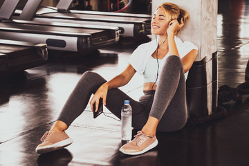 Young happy woman sitting and listening to music by smartphone while having exercise break at the gym.