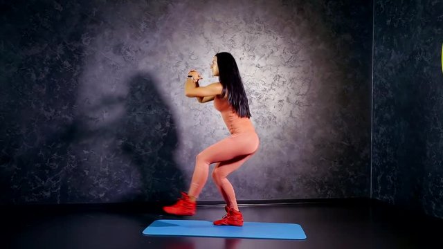 young girl trainer in a sports jumpsuit shows exercise is the squat with the single leg raise in the horizon and touch the fingers of the hand