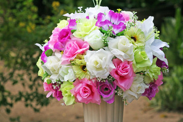 Artificial muticolored rose flowers in white vase on green garden background