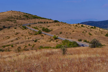 The asphalted road snakes along the slope overgrown with dry grass sunburned in the sun. The car is driving along the road towards the sun shining in his face.