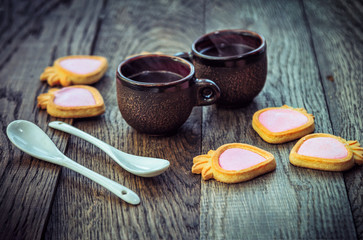 Ceramic cups with hot freshly brewed coffee, strawberry shape cookies with pink cream and white spoons on a wooden table. Sweet snack or breakfast.