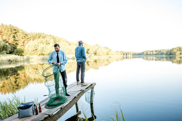 Naklejka premium Two male friends preparing for fishing standing with fishing net and rod on the wooden pier during the morning light on the lake