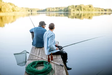 Crédence de cuisine en verre imprimé Pêcher Deux amis masculins vêtus de chemises bleues pêchant avec filet et canne assis sur la jetée en bois pendant la lumière du matin sur le lac
