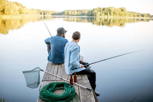 Two male friends dressed in blue shirts fishing together with net and rod sitting on the wooden pier during the morning light on the lake
