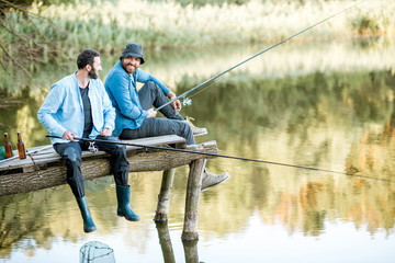 Zwei männliche Freunde in blauen Hemden, die zusammen mit Netz und Rute angeln, sitzen auf dem Holzsteg während des Morgenlichts auf dem See