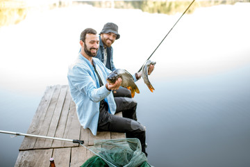 Two happy fishermen holding caught fish sitting on the wooden pier during the fishing on the lake at the morning