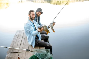 Foto op Plexiglas Vissen Twee gelukkige vissers die gevangen vis vasthouden terwijl ze op de houten pier zitten tijdens het vissen op het meer in de ochtend