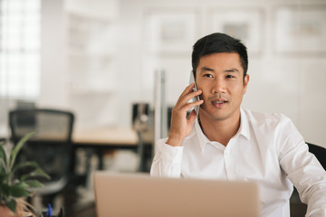 Asian businessman sitting at his desk talking on a cellphone