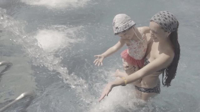 A woman and her daughter are having fun playing with water in the pool. A little girl is catching the streams of water. S-log, ungraded