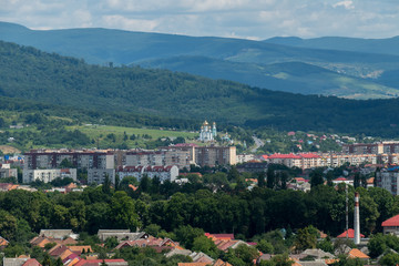 Fototapeta premium Residential houses and a church with golden domes located at the foot of wide massive mountains with green forests