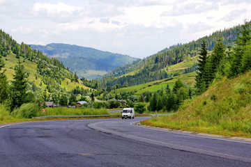 The car rides between the high green mountain slopes along the road winding between them. With the roofs of rural houses seen among the trees standing on the roadside.