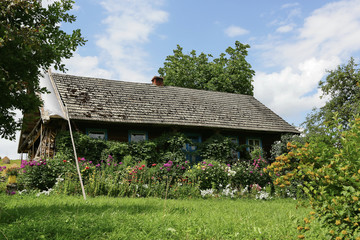 A wooden country cottage is covered with tiled windows with blue windows in an oasis of green trees.