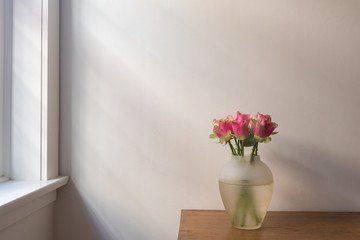 Pink roses in glass vase on wooden table next to window with soft shadows (selective focus)