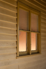 A window on the side of a wooden house in the evening with a softly glowing orange interior seen from the outside