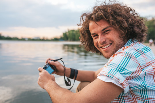 Rear view of happy handsome male with curly hair taking photos of nature on his digital camera. Young smiling man wears casual shirt, with digital camera posing in the park. People, travel, lifestyle