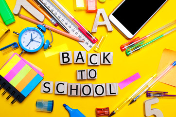 inscription from wooden cubes "back to school". school accessories on a desk on a yellow background. concept of education. stationery. watches, colored pens, phone, markers. notebook.
