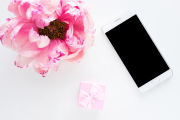 White background with copy space, smart phone lying on the table, pink peony flower, small gift for girl, advertising space, top view