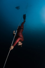 Freediver in red wetsuit descends along the rope. Safety freediver follows him on the background.