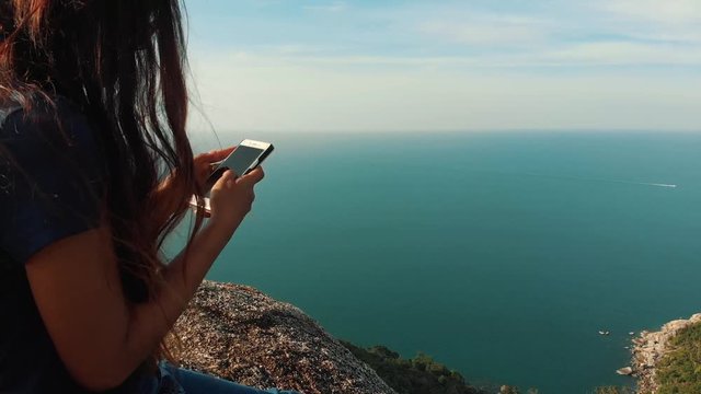Cute Asian girl uses a mobile phone sitting on a cliff face smiling Blue sea and green jungle on a background. Woman with smartphone sitting on the mountain. Asia Thailand