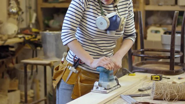 Tracking shot of young woman with respirator using belt sander on wooden plank in carpentry workshop