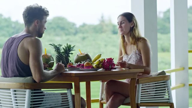 Medium shot of Caucasian couple sitting opposite each other at table on balcony and having eating tropical fruits for breakfast