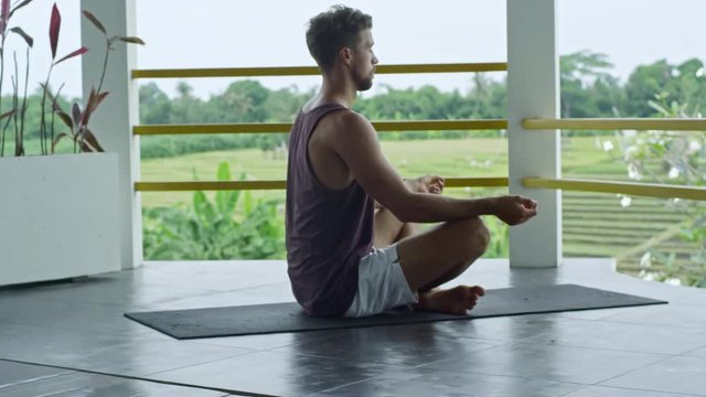 Relaxed Caucasian man sitting on yoga mat and doing meditation practice in lotus position when enjoying fresh air on balcony