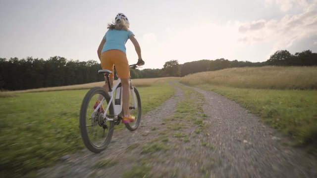 4K sport footage, 44 years old fit woman cycling up gravel road between high dry grass and meadow with electric mountain bike in  summer sunny back light
