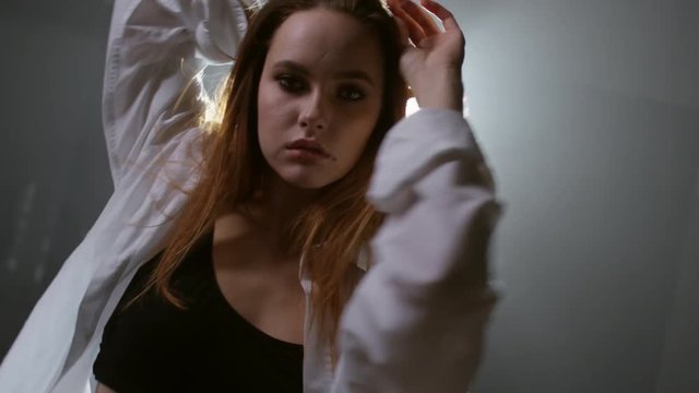 Medium shot of sensual young woman in top and mens shirt touching her hair and moving seductively before camera in dark foggy studio with bright spotlight