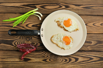 Two fried eggs in metal beige pan beside green fresh onion and dried red hot peppers on old wooden brown table. Top view