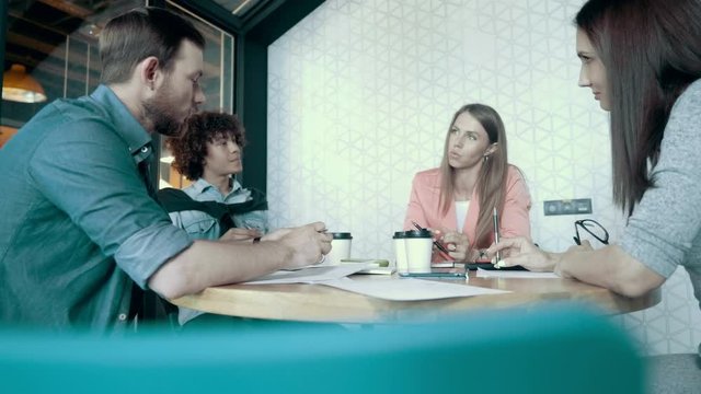 European woman emotionally communicates with team members actively gestures african man drinking coffee multiethnic creative business team meeting brainstorming in modern start up office dolly shot