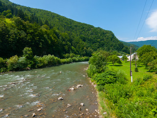 The green slopes of the mountains are overgrown with dense forest with the current shallow river with rocks on the bottom and green grass on the opposite shore.