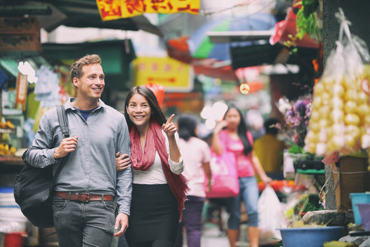 Interracial Couple Tourists Walking Shopping In Chinese Market In Hong Kong, China. Young People Traveling In Asia Looking At Local Street Food. Asian Woman, Caucasian Man.