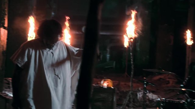 Spooky psycho in a mask and a white hoodie plays drums with a long haired drummer in an abandoned factory with lighted torches on background