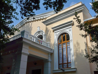 The facade of a beautiful building with a tall window and stucco on the wall with a balcony with white rails and trees growing next to it.