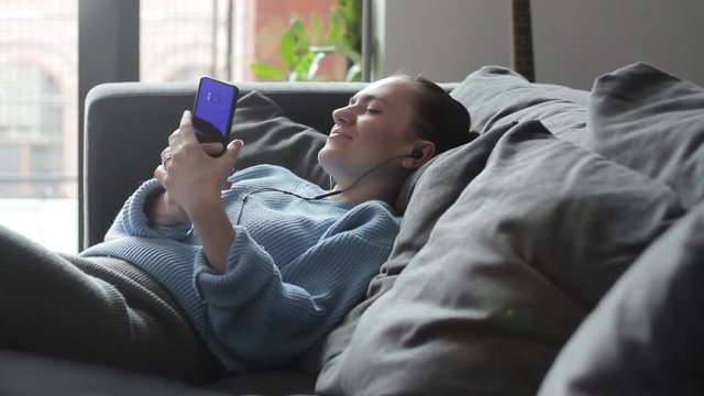Smiling pretty woman in headphones using smartphone while sitting on a sofa at home
