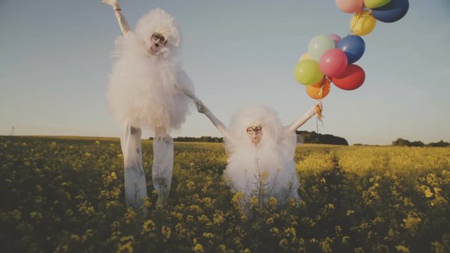 Two funny clowns mime in white air suits on stilts and with colored balloons walk around the field and have fun.