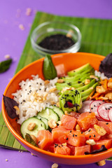 Hawaiian salad Poke with salmon fish, avocado, Korean carrots, cucumbers, radish and rice, with black sesame. in an orange plate. On the purple trend background of 2018. copy space, top view