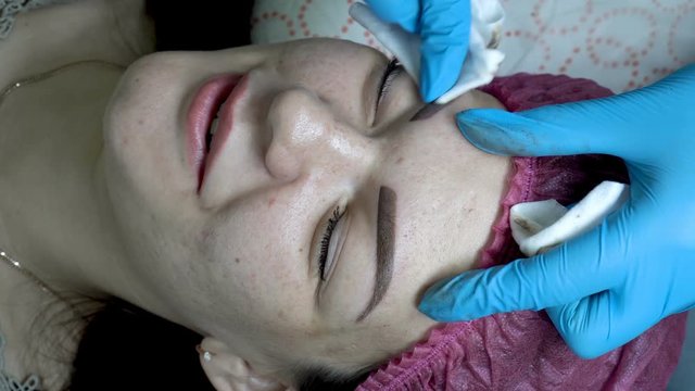 The beautician treats the skin around the eyebrows of the girl in the beauty salon after the procedure of permanent makeup eyebrows. Extremely close-up. The view from the top. 4K, 25 fps.
