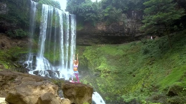 lady holds yoga pose with hands above head by waterfall