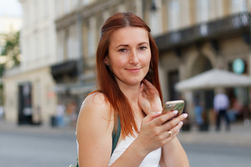 Young beautiful redhead woman touching her neck, holding smartphone and walking down a street