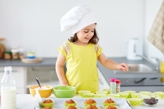 family, cooking and people concept - little girl in chefs toque baking muffins or cupcakes with sprinkles at home
