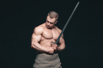 Bodybuilder man posing with a sword isolated on black background. Serious shirtless man...